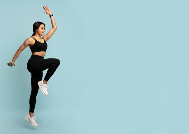 Fitness-Konzept. Fit junge afrikanisch-amerikanische Frau springt, hebt das Bein, blauer Hintergrund mit leerem Raum - Foto, Bild