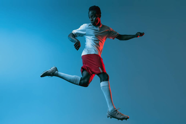 Nahaufnahme afrikanischer Fußballspieler, der isoliert auf blauem Hintergrund im Neonlicht springt - Foto, Bild