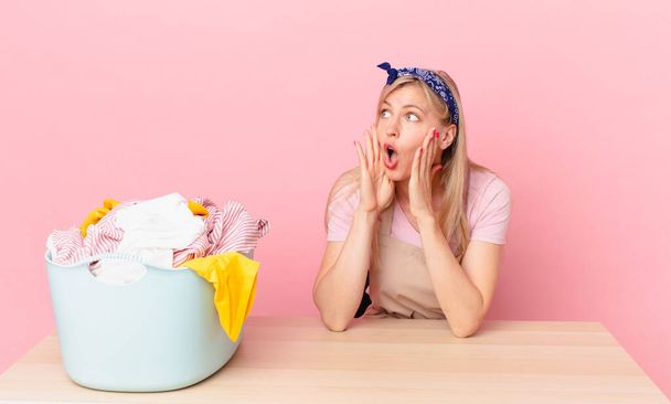 young blonde woman feeling happy, excited and surprised. wash clothes concept - Фото, изображение