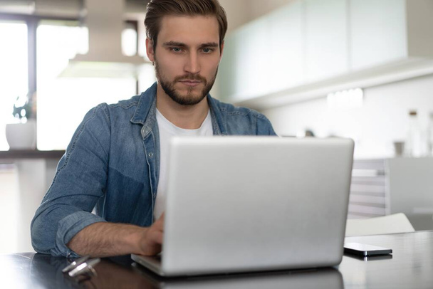Young casual man working on laptop online, sitting at table in kitchen, looking at computer screen - Photo, Image