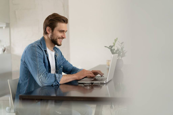 Mladý ležérní muž pracující na notebooku on-line, sedí u stolu v kuchyni, dívá se na obrazovku počítače - Fotografie, Obrázek