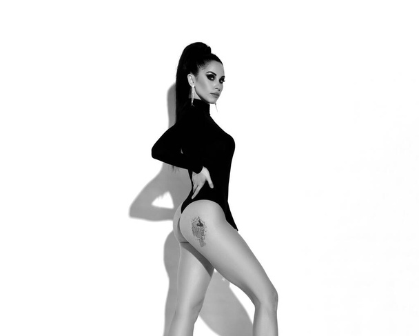 Черно-белый портрет сексуальной стройненькой брюнетки в черном теле, стоящей боком с татуировкой на ягодицах - Фото, изображение