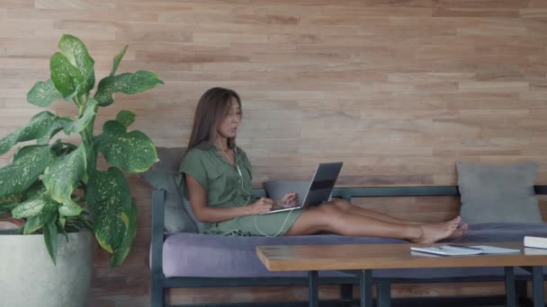 Wide shot of young happy Caucasian woman wearing wired headset, sitting on sofa with feet up by large plant in pot, having video conversation on portable computer on her lap - Footage, Video