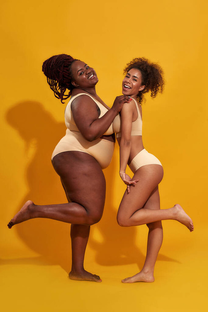 Two gorgeous young african american women in underwear with different body size lifting leg while posing together isolated over yellow background - Fotó, kép