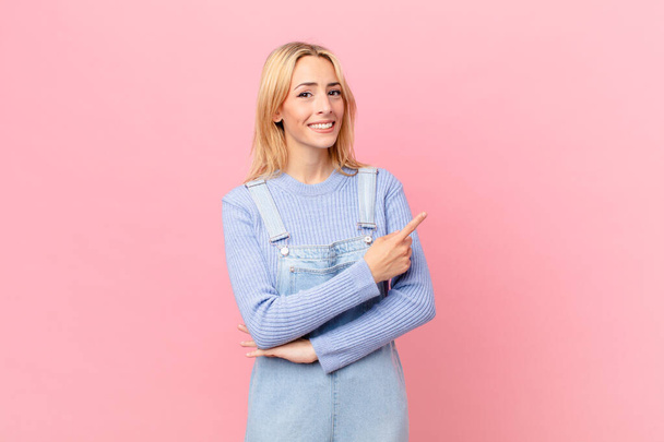 young blonde woman smiling cheerfully, feeling happy and pointing to the side - Foto, afbeelding