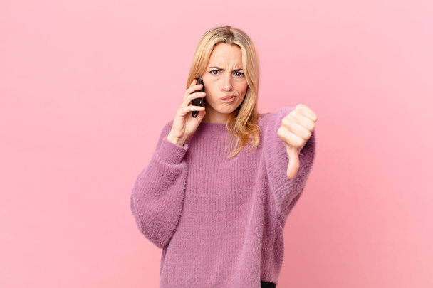 young blonde woman feeling cross,showing thumbs down and talking with a smartphone - Foto, Bild