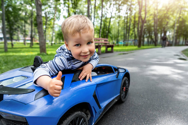 Portrét roztomilého malého kavkazského blonďatého batolete chlapce baví jezdit na elektricky poháněné autíčko asfaltovou cestou road city park v letní den. Happy child show thumbs up gesture playing outdoors - Fotografie, Obrázek