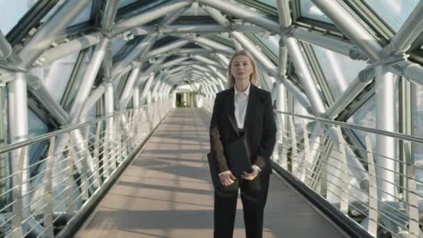 Средний медлительный портрет успешной деловой женщины среднего возраста, держащей кожаную папку, смотрящей на камеру, стоящую на крытом стеклянном мосту офисного здания высокого класса - Кадры, видео