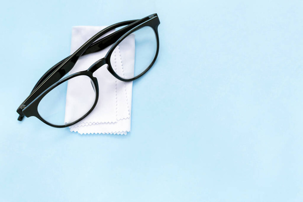 Black rimmed glasses and cleaning cloth on blue background with copy space. White soft tissue for wiping eyeglasses from dirt and dust. Vision correction, optics care, ophthalmology, optometry. - Фото, изображение