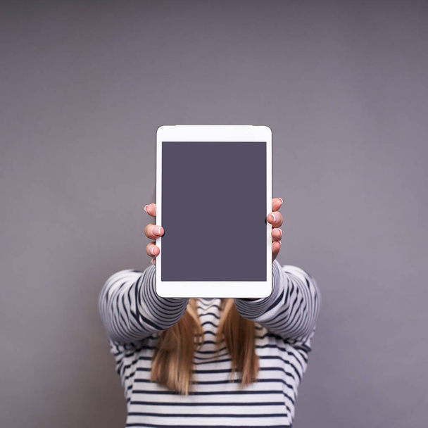 Vertical shot, close-up, copy space on her digital screen. Fashionable young woman, in a striped sweater, holds a tablet at face level, posing against a gray background. No face concept. Copy space. - Photo, Image