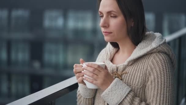 Blanke vrouw verblijft op het balkon tijdens de sneeuwval met een kopje warme koffie of thee. Ze kijkt naar de sneeuwvlokken en ademt de ijzige frisse lucht in. - Video