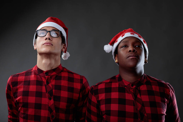 Dos amigos de los hombres que usan los sombreros de Navidad de Santa Claus y las camisas rojas a cuadros que emparejan de pie lado a lado mirando para arriba con expresiones serias contra un fondo gris del estudio en un concepto del día de fiesta - Foto, Imagen