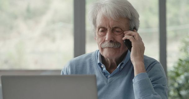 Happy senior man sitting at desk and receiving a phone call on his smartphone - Foto, Imagem