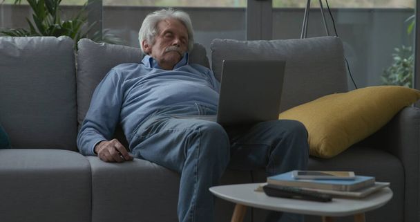 Elderly man falling asleep on the couch while watching videos on his laptop - Φωτογραφία, εικόνα