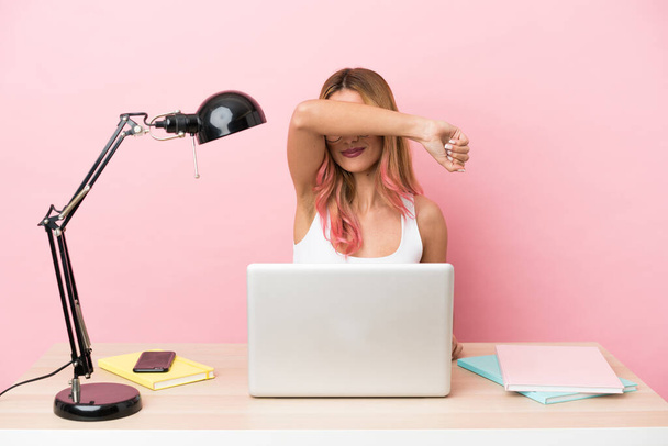 Giovane studentessa in un posto di lavoro con un computer portatile su sfondo rosa che copre gli occhi dalle mani - Foto, immagini