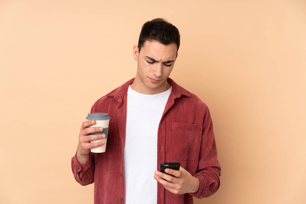 Mladý kavkazský pohledný muž izolovaný na béžovém pozadí držící kávu odnést a mobilní - Fotografie, Obrázek