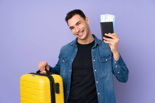 Beyaz tenli yakışıklı, mor arka planda izole edilmiş, bavul ve pasaportla tatilde. - Fotoğraf, Görsel