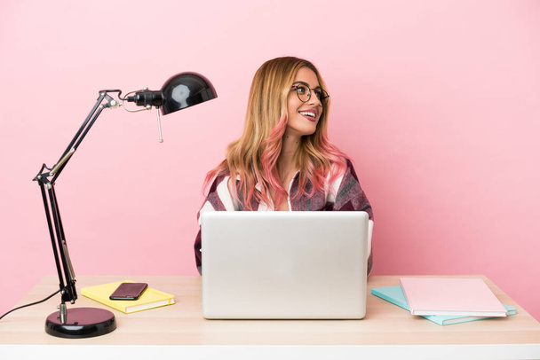 Junge Studentin an einem Arbeitsplatz mit Laptop vor rosa Hintergrund, die Arme verschränkt und glücklich - Foto, Bild