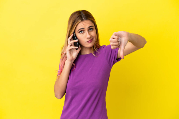 İzole edilmiş sarı arka planda cep telefonu kullanan genç bir kadın, olumsuz ifade ile baş parmağını aşağı gösteriyor. - Fotoğraf, Görsel