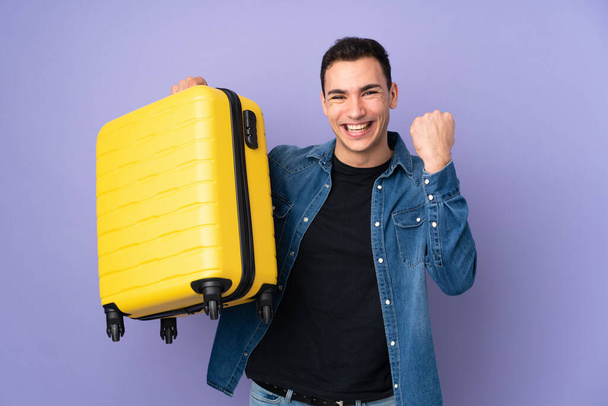 Jeune homme beau caucasien isolé sur fond violet en vacances avec valise de voyage - Photo, image