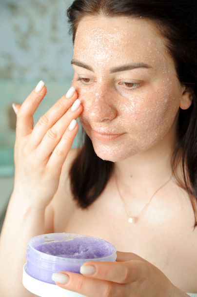 Mooie jonge vrouw brengt violette scrub aan op haar gezicht. Vrouw doet schoonheidsbehandelingen thuis.  - Foto, afbeelding