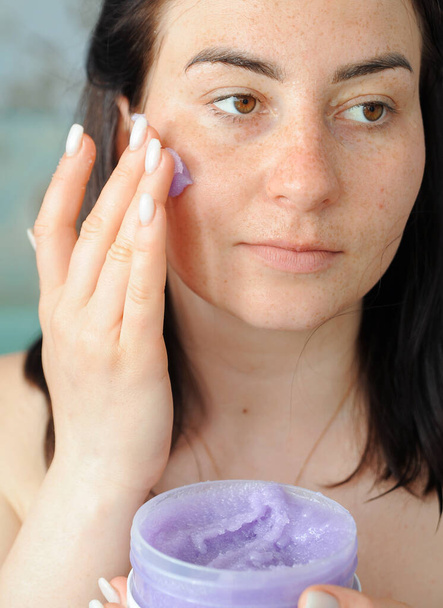 Mulher bonita aplica esfoliação violeta em seu rosto. Mulher fazendo tratamentos de beleza em casa.  - Foto, Imagem