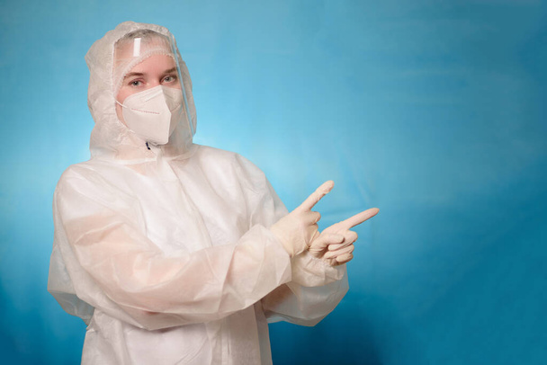 Un medico in una tuta protettiva contro il Covid-19 coronavirus su uno sfondo blu punta le dita nello spazio di copia - Foto, immagini