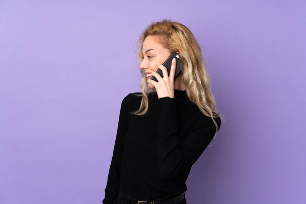 Mujer rubia joven aislada sobre fondo morado manteniendo una conversación con el teléfono móvil con alguien - Foto, imagen