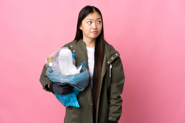 Kiinalainen tyttö tilalla laukku täynnä muovipulloja kierrättää yli eristetty vaaleanpunainen tausta ottaa epäilyksiä etsiessään - Valokuva, kuva