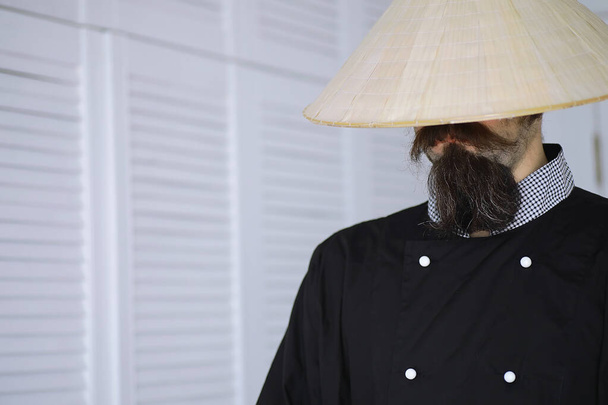 Μια ξεκαρδιστική παρωδία ενός Ασιάτη με βιετναμέζικο καπέλο και μούσι. Πορτρέτο. Ασιάτης σεφ καφέ. - Φωτογραφία, εικόνα