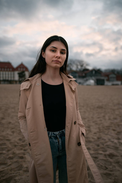 Lifestyle portrait of brunette wears trench coat, black shirt and jeans on the ocean coast. Girl posing near classical hotel by the seaside during cold time of the year. - Φωτογραφία, εικόνα