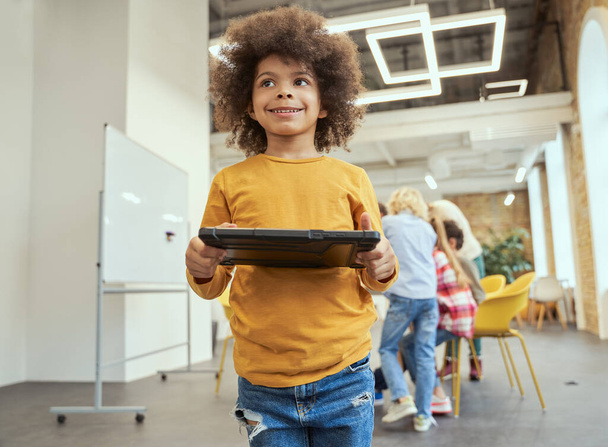 Portrait of sweet little boy with afro hair looking away, holding tablet pc, standing in a classroom during STEM lesson - Zdjęcie, obraz