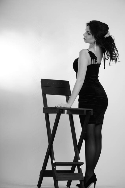 sexy beautiful brunette woman in little black dress and tights with high heels posing with director studio high chair isolated on white background  - Photo, Image