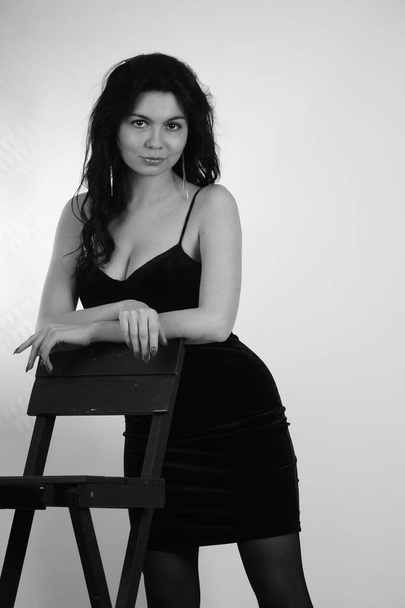 σέξι όμορφη μελαχρινή γυναίκα σε λίγο μαύρο φόρεμα και καλσόν με ψηλά τακούνια ποζάρουν με στούντιο σκηνοθέτη ψηλή καρέκλα απομονωμένη σε λευκό φόντο  - Φωτογραφία, εικόνα