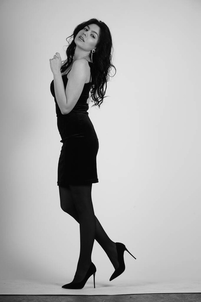 сексуальна красива брюнетка в маленькій чорній сукні і колготках з високими підборами позує в студії на білому тлі
  - Фото, зображення