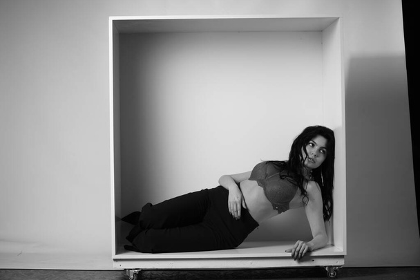 сексуальная красивая брюнетка женщина в нижнем белье и модные брюки колокольчика позирует в декоративной коробке в студии на белом фоне  - Фото, изображение