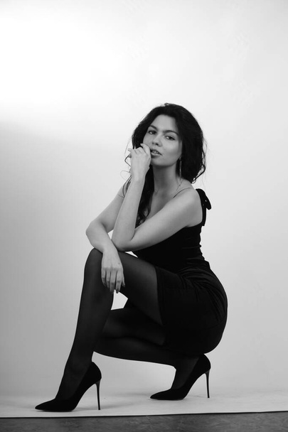 σέξι όμορφη μελαχρινή γυναίκα σε λίγο μαύρο φόρεμα και καλσόν με ψηλά τακούνια ποζάρουν στο στούντιο σε λευκό φόντο  - Φωτογραφία, εικόνα
