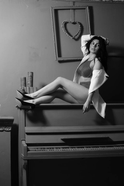 gyönyörű barna nő fehérnemű és fehér blézer pózol zongorán a stúdióban  - Fotó, kép