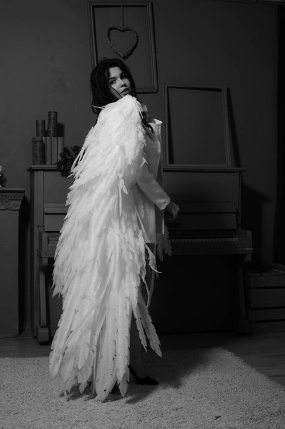 mooie brunette vrouw in blazer en witte engel vleugels poseren in de buurt van piano in studio  - Foto, afbeelding