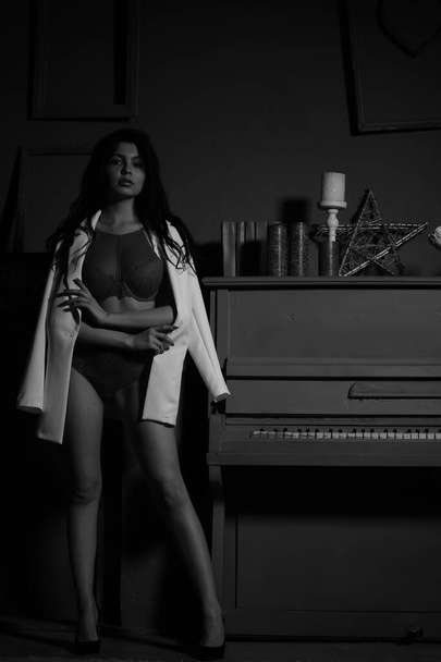 bella donna bruna in lingerie e blazer bianco in posa vicino al pianoforte in studio  - Foto, immagini