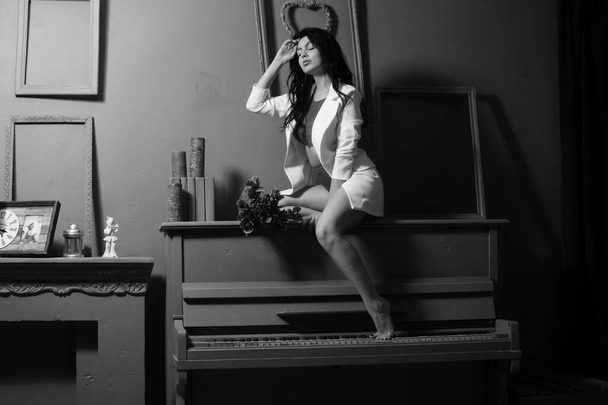 İç çamaşırlı, beyaz ceketli güzel esmer kadın. Stüdyoda piyanonun üzerinde çiçek demeti poz veriyor.  - Fotoğraf, Görsel
