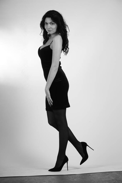 sexy schöne brünette Frau in kleinem schwarzen Kleid und Strumpfhosen mit High Heels posiert im Studio auf weißem Hintergrund  - Foto, Bild