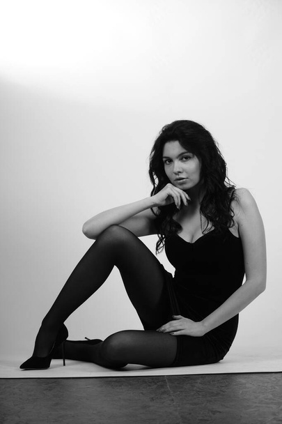 sexy schöne brünette Frau in kleinem schwarzen Kleid und Strumpfhosen mit High Heels posiert im Studio auf weißem Hintergrund  - Foto, Bild