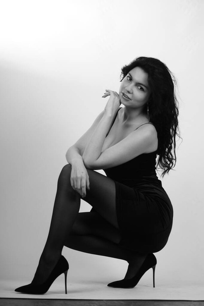 сексуальная красивая брюнетка женщина в маленьком черном платье и колготки с высокими каблуками позирует в студии на белом фоне  - Фото, изображение