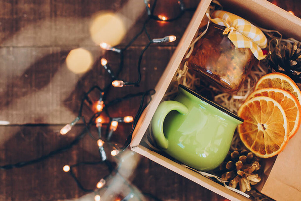 Caja de regalo con juego de té. Té, taza, mermelada o miel, especias, naranjas secas, anís en caja en mesa de madera con luces de Navidad, vista superior. Hermoso regalo, paquete de cuidado para la familia y amigos - Foto, imagen