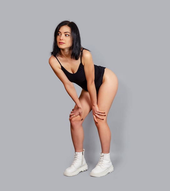 Fitness woman studio portrait in black bodysuit, white sneakers, half-bent, leaning on knees with both hands, ass back, looking at camera, on gray background. - 写真・画像