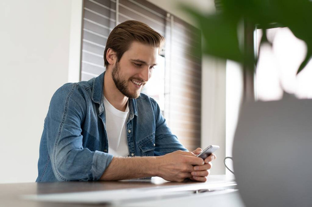 Confident man holding smartphone, sitting in kitchen, chatting online with friends or colleagues, browsing app - Photo, Image