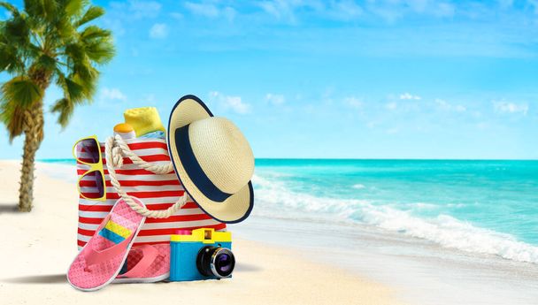 Bolsa de playa de verano y accesorios sombrero de paja, chanclas y gafas de sol en la playa de arena y el mar azul en el fondo - Foto, imagen