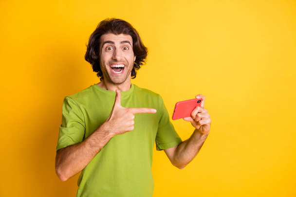 Photo of crazy excited guy hold telephone indicate finger wear green t-shirt isolated yellow color background - Photo, image