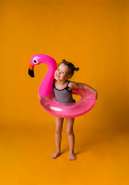 vrolijk klein meisje in een gestreept zwempak en met een opblaasbare zwemcirkel in de vorm van een flamingo staat op een gele achtergrond met een plek voor tekst. Zomervakantie - Foto, afbeelding